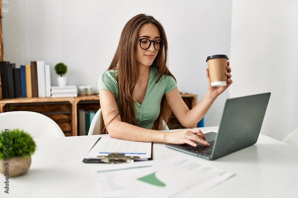 Young hispanic woman using laptop drinking coffee sitting on table at home Young hispanic woman using laptop drinking coffee sitting on table at home