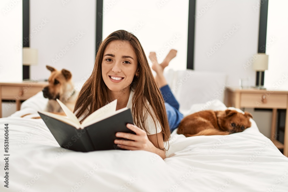 Young hispanic woman reading book lying on bed with dogs at bedroom Young hispanic woman reading book lying on bed with dogs at bedroom