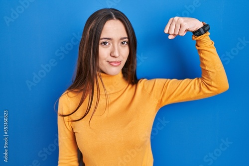 Young brunette woman standing over blue background strong person showing arm muscle, confident and proud of power