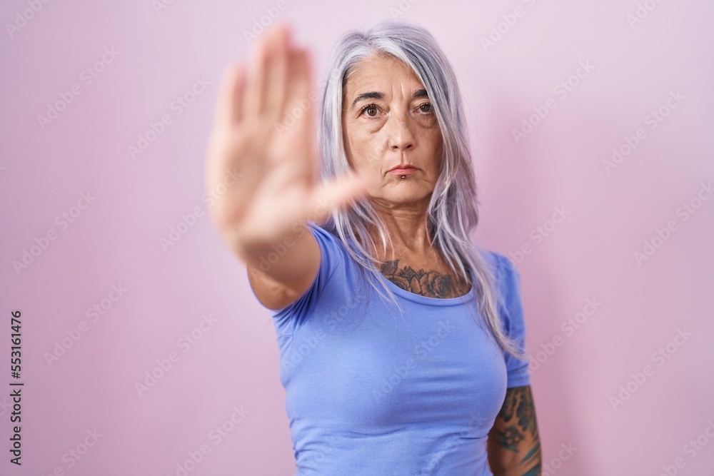 Middle age woman with tattoos standing over pink background doing stop sing with palm of the hand. warning expression with negative and serious gesture on the face.