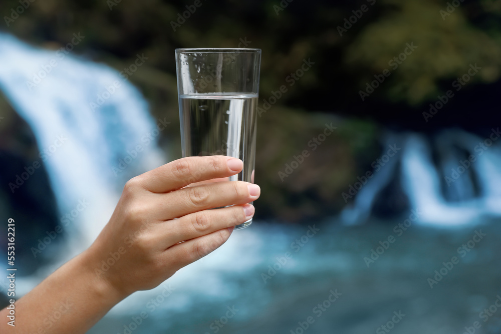 Obraz premium Woman holding glass of fresh water near waterfall outdoors, closeup. Space for text