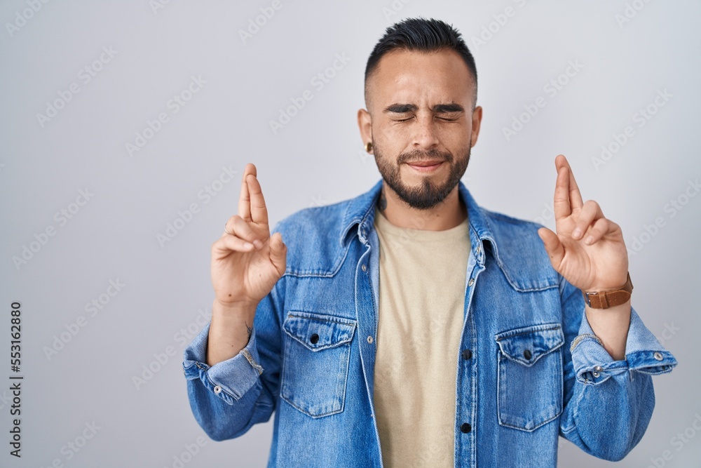 Young hispanic man standing over isolated background gesturing finger crossed smiling with hope and eyes closed. luck and superstitious concept. Young hispanic man standing over isolated background gesturing finger crossed smiling with hope and eyes closed. luck and superstitious concept.