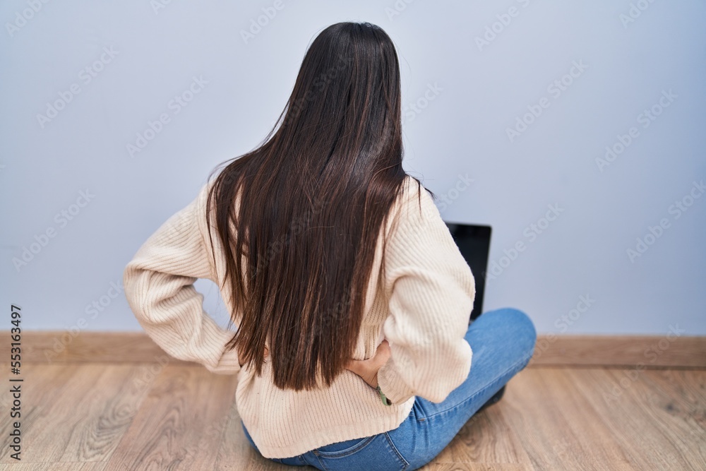 Young woman using laptop sitting on the floor at home standing backwards looking away with arms on body Young woman using laptop sitting on the floor at home standing backwards looking away with arms on body