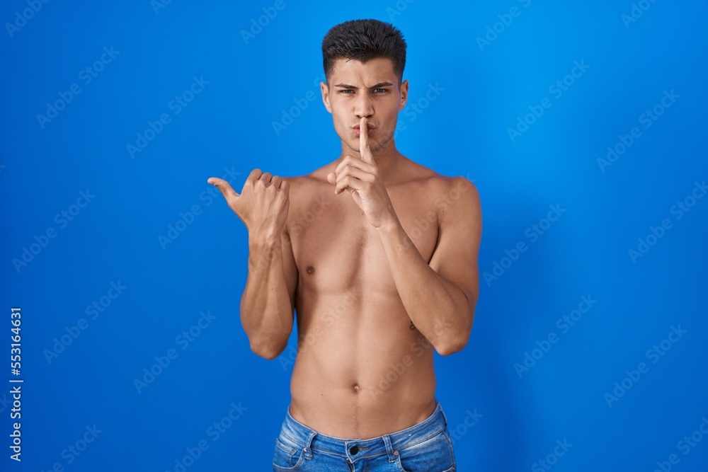 Young hispanic man standing shirtless over blue background asking to be quiet with finger on lips pointing with hand to the side. silence and secret concept.