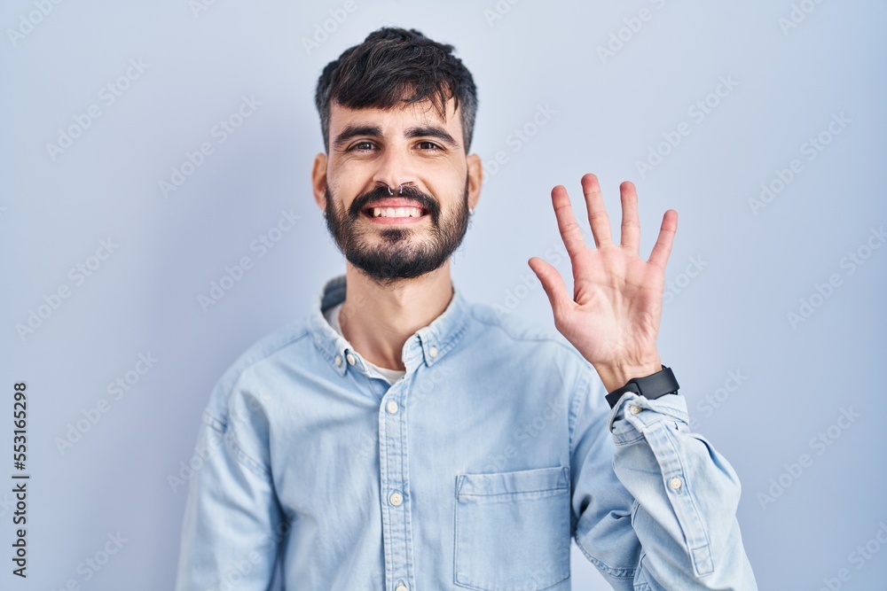 Young hispanic man with beard standing over blue background showing and pointing up with fingers number five while smiling confident and happy. Young hispanic man with beard standing over blue background showing and pointing up with fingers number five while smiling confident and happy.