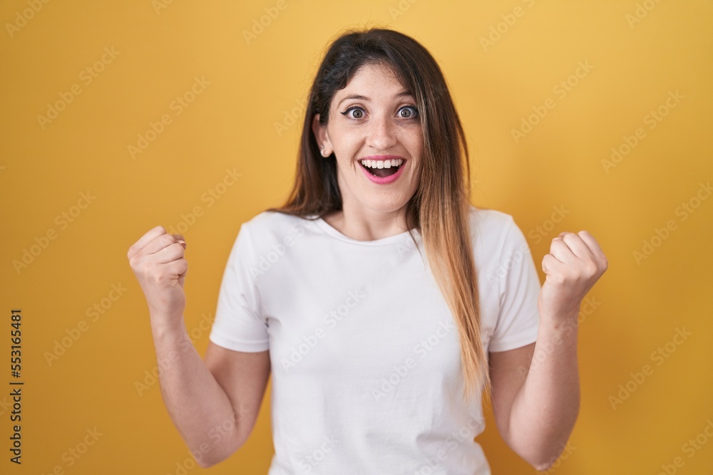 Young brunette woman standing over yellow background celebrating surprised and amazed for success with arms raised and open eyes. winner concept. Young brunette woman standing over yellow background celebrating surprised and amazed for success with arms raised and open eyes. winner concept.