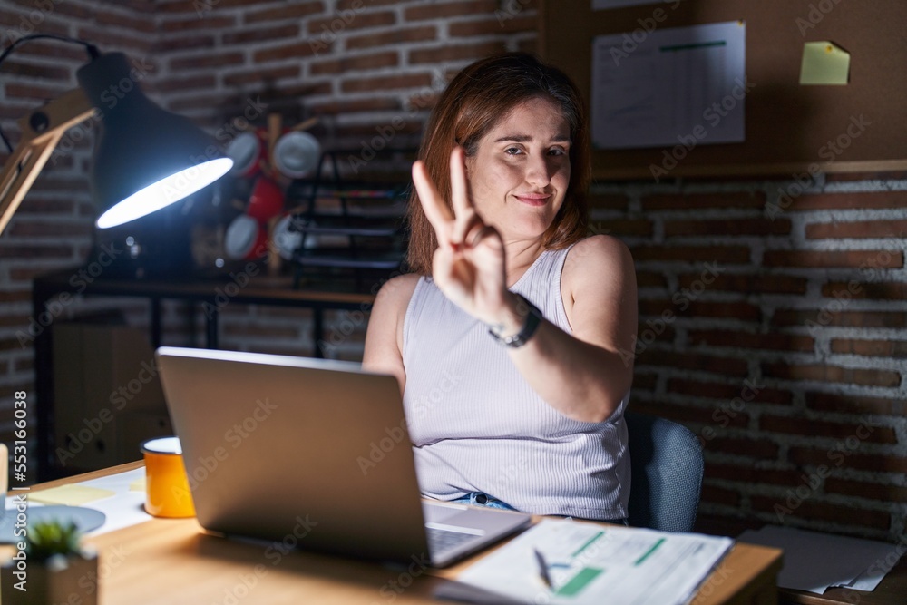 Brunette woman working at the office at night smiling looking to the camera showing fingers doing victory sign. number two. Brunette woman working at the office at night smiling looking to the camera showing fingers doing victory sign. number two.