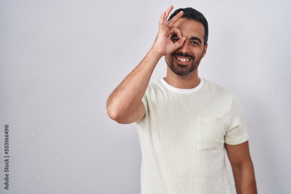 Hispanic man with beard standing over isolated background doing ok gesture with hand smiling, eye looking through fingers with happy face. Hispanic man with beard standing over isolated background doing ok gesture with hand smiling, eye looking through fingers with happy face.