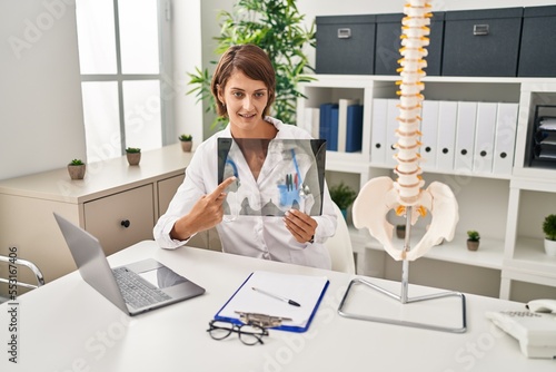 Young beautiful hispanic woman doctor pointing with finger to vertebral column xray at clinic photo