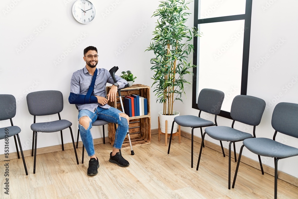 Young arab man smiling confident sitting on chair with arm sling holding crutch at clinic waiting room Young arab man smiling confident sitting on chair with arm sling holding crutch at clinic waiting room