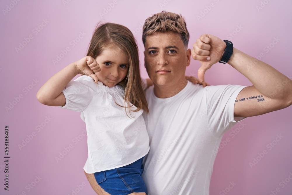 Young father hugging daughter over pink background with angry face, negative sign showing dislike with thumbs down, rejection concept Young father hugging daughter over pink background with angry face, negative sign showing dislike with thumbs down, rejection concept
