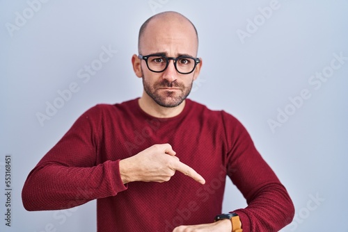 Young bald man with beard standing over white background wearing glasses in hurry pointing to watch time, impatience, upset and angry for deadline delay