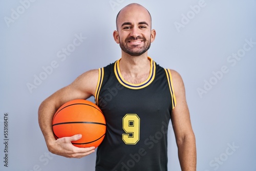 Young bald man with beard wearing basketball uniform holding ball with a happy and cool smile on face. lucky person.