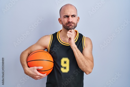 Young bald man with beard wearing basketball uniform holding ball thinking concentrated about doubt with finger on chin and looking up wondering