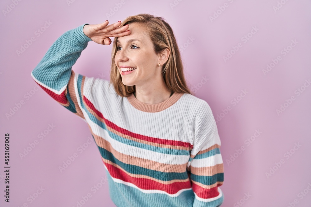 Young blonde woman standing over pink background very happy and smiling looking far away with hand over head. searching concept. Young blonde woman standing over pink background very happy and smiling looking far away with hand over head. searching concept.
