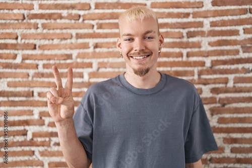 Young caucasian man standing over bricks wall showing and pointing up with fingers number two while smiling confident and happy.