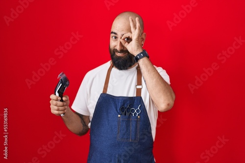 Young hispanic man with beard and tattoos wearing barber apron holding razor doing ok gesture with hand smiling, eye looking through fingers with happy face. © Krakenimages.com