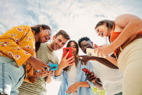 Slika na platnu Low angle view of a group of smiling multiracial teenagers addicted to smartphones, watching funny videos, shopping online, enjoying outdoors