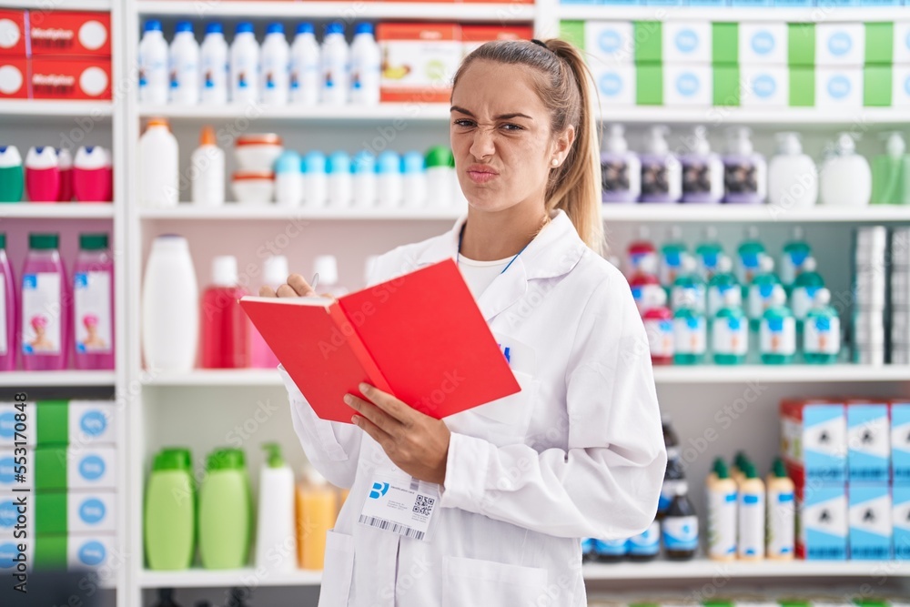 Young blonde woman working at pharmacy drugstore holding notebook skeptic and nervous, frowning upset because of problem. negative person.