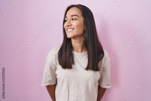 Young hispanic woman standing over pink background looking away to side with smile on face, natural expression. laughing confident.