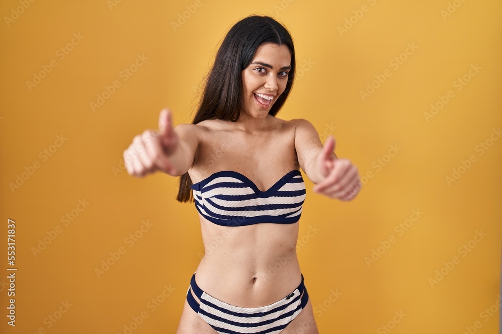 Naklejka premium Young brunette woman wearing bikini over yellow background approving doing positive gesture with hand, thumbs up smiling and happy for success. winner gesture.