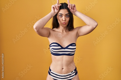 Young brunette woman wearing bikini over yellow background doing funny gesture with finger over head as bull horns