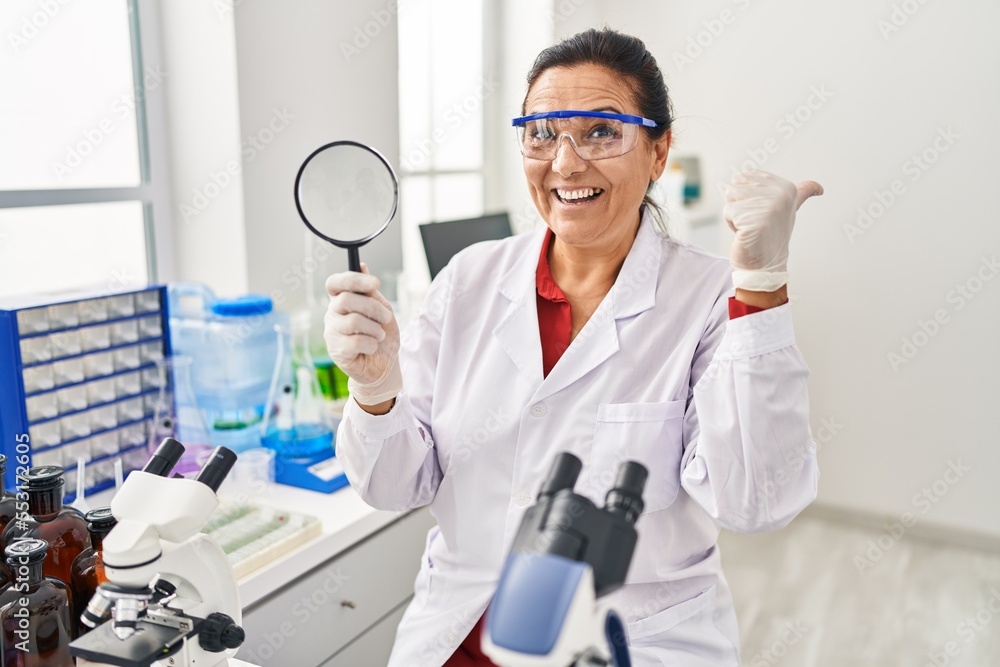 Middle age hispanic woman working at scientist laboratory holding magnifying glass pointing thumb up to the side smiling happy with open mouth Middle age hispanic woman working at scientist laboratory holding magnifying glass pointing thumb up to the side smiling happy with open mouth
