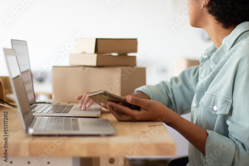 Young online shop worker holding cellphone and checking order details on laptop. Black woman working on computer surrounded by cardboard parcels. Startup business, e-commerce concept © KAMPUS