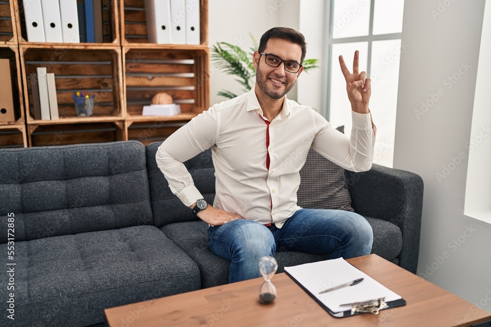 Young hispanic man with beard working at consultation office smiling looking to the camera showing fingers doing victory sign. number two. Young hispanic man with beard working at consultation office smiling looking to the camera showing fingers doing victory sign. number two.