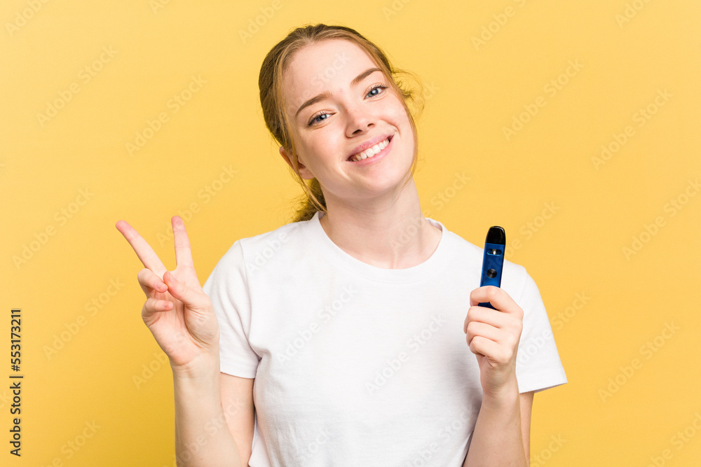 Young cute woman holding a vaper isolated on yellow background joyful and carefree showing a peace symbol with fingers. Young cute woman holding a vaper isolated on yellow background joyful and carefree showing a peace symbol with fingers.