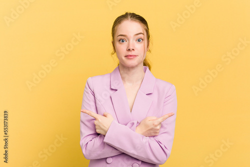 Young caucasian redhead woman isolated on yellow background points sideways, is trying to choose between two options.