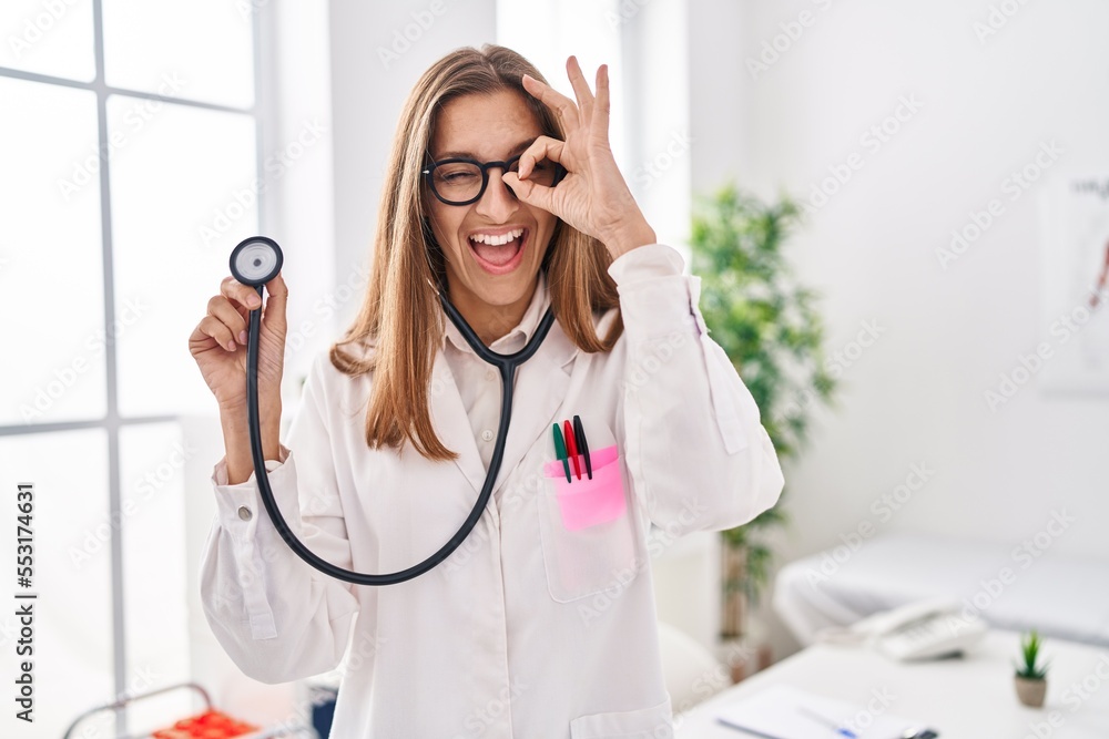 Young woman wearing doctor uniform holding stethoscope smiling happy doing ok sign with hand on eye looking through fingers Young woman wearing doctor uniform holding stethoscope smiling happy doing ok sign with hand on eye looking through fingers
