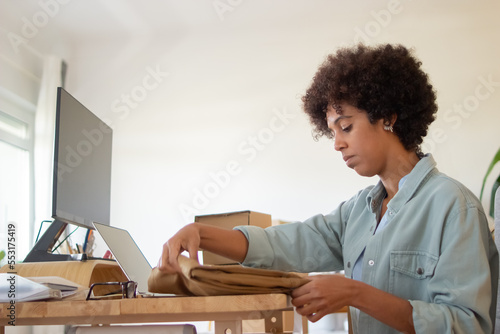 Serious blackwoman folding garment for dispatch. Concentrated African American online store employeer working in office. Packing package concept photo