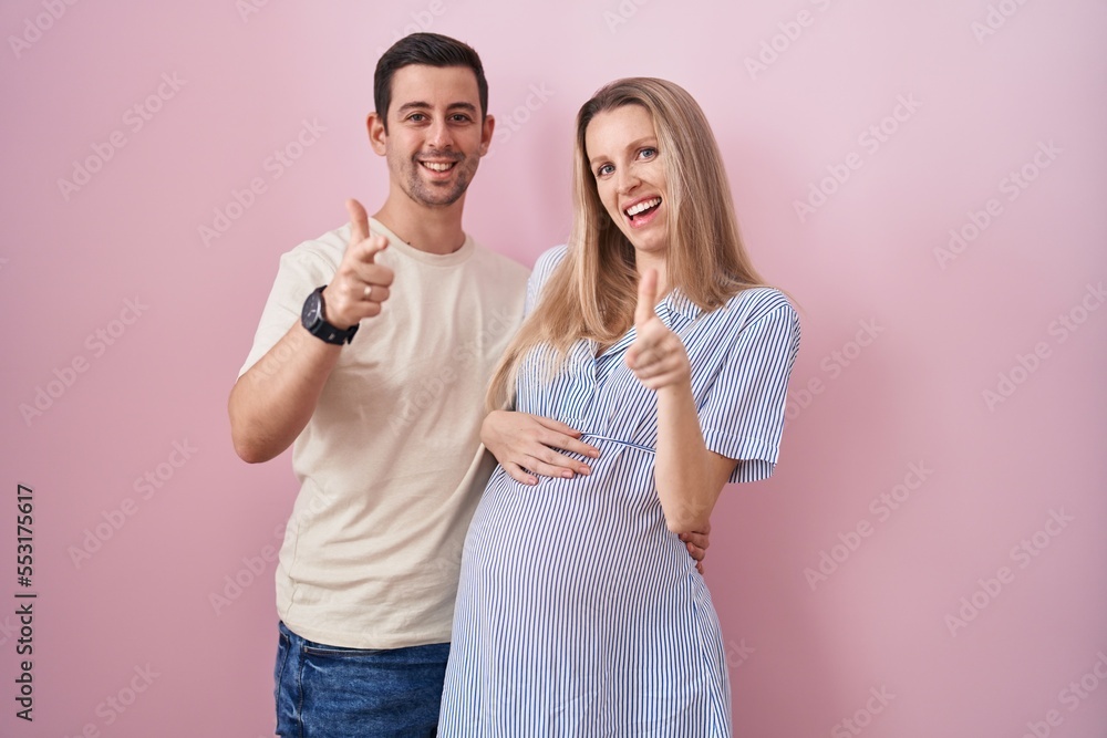 Young couple expecting a baby standing over pink background pointing fingers to camera with happy and funny face. good energy and vibes. Young couple expecting a baby standing over pink background pointing fingers to camera with happy and funny face. good energy and vibes.