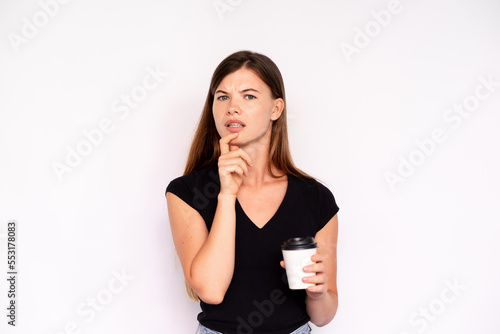 Portrait of incredulous young woman holding coffee over white background. Caucasian lady wearing black T-shirt looking at camera with doubtful expression. Doubt concept photo