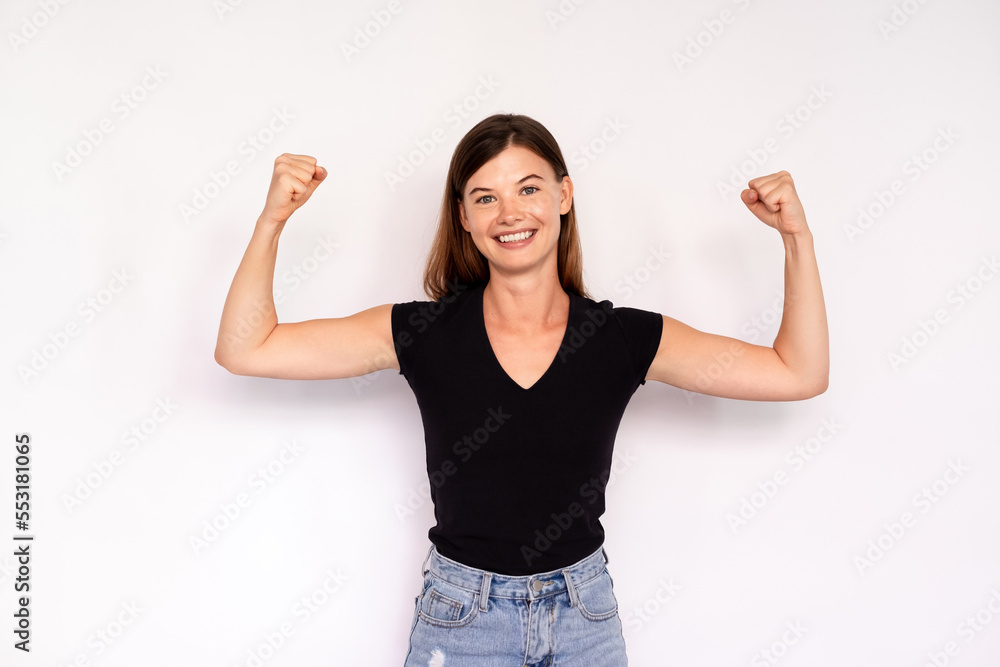 Portrait of happy young woman showing muscles over white background. Caucasian lady wearing black T-shirt and jeans looking at camera and smiling. Strength and fitness concept Portrait of happy young woman showing muscles over white background. Caucasian lady wearing black T-shirt and jeans looking at camera and smiling. Strength and fitness concept