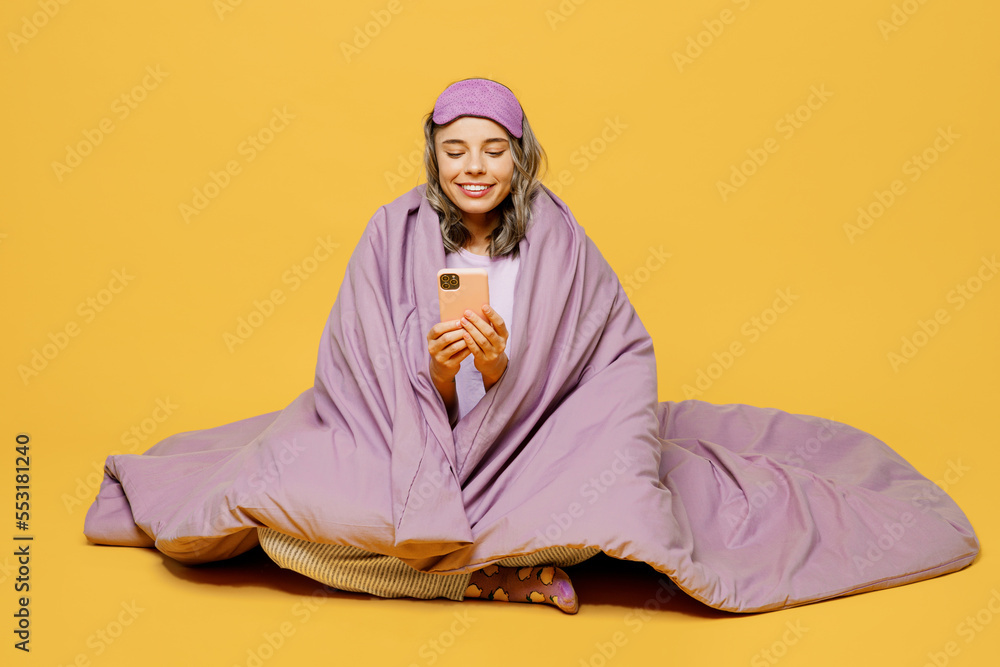 Fototapeta premium Full body young woman she wears purple pyjamas jam sleep eye mask rest relax at home sit wrap covered blanket duvet hold use mobile cell phone isolated on plain yellow background. Night nap concept.