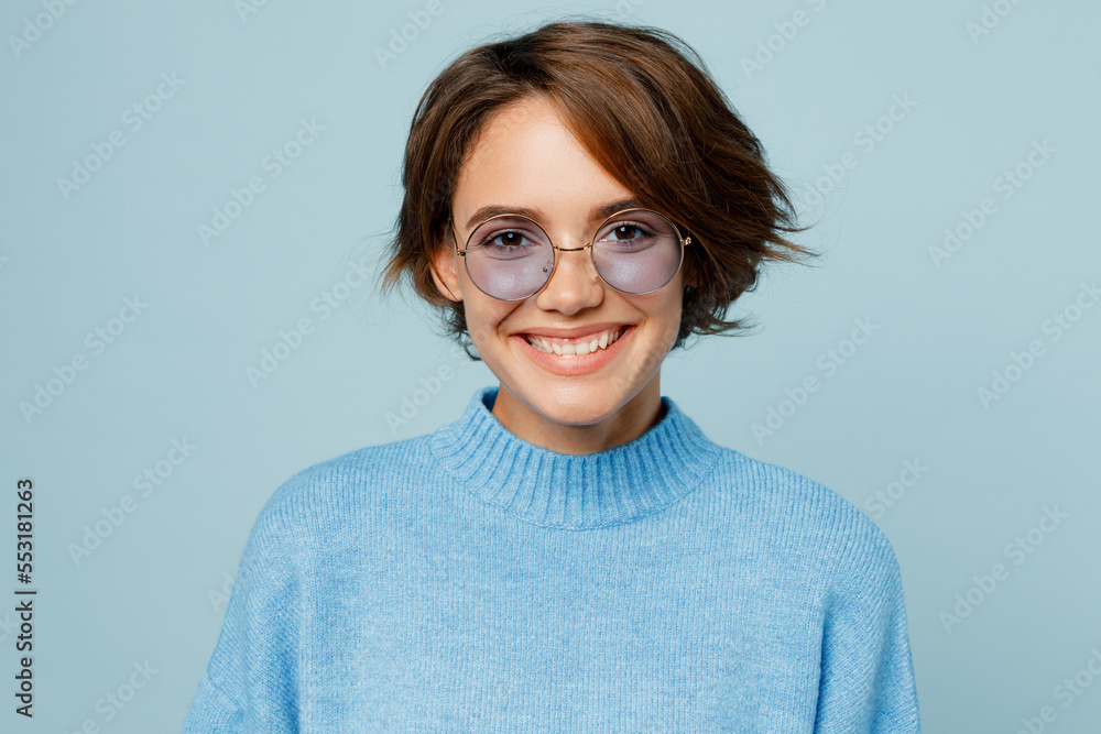 Young smiling happy cheerful fashionable fun caucasian woman wearing knitted sweater glasses look camera isolated on plain pastel light blue cyan background studio portrait. People lifestyle concept.