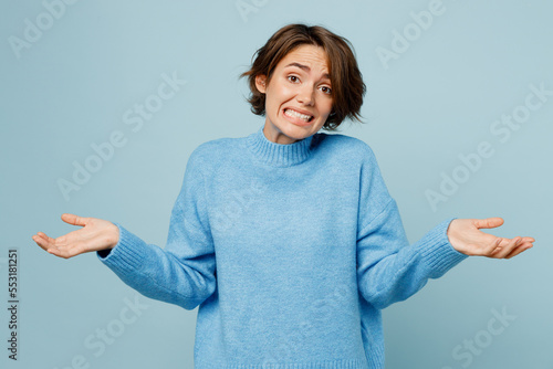 Young caucasian woman wear knitted sweater shrugging shoulders looking puzzled, have no idea spread hands isolated on plain pastel light blue cyan background studio portrait. People lifestyle concept. photo