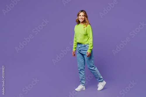 Full body side view smiling fun young woman 30s she wearing casual green knitted sweater walking going look camera isolated on plain pastel purple background studio portrait. People lifestyle concept.