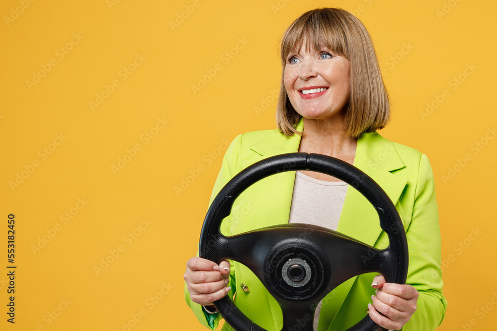 Elderly smiling woman 50s years old wearing green jacket white t-shirt hold steering wheel driving car look aside on area isolated on plain yellow background studio portrait. People lifestyle concept. Elderly smiling woman 50s years old wearing green jacket white t-shirt hold steering wheel driving car look aside on area isolated on plain yellow background studio portrait. People lifestyle concept.