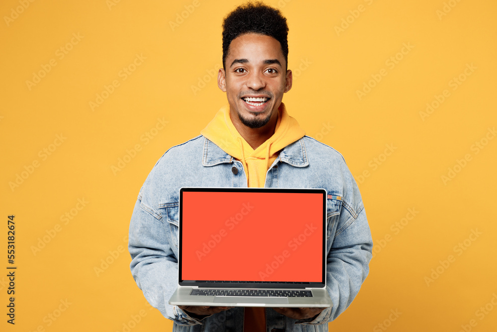 Young fun smart IT man of African American ethnicity in denim jacket hoody look camera hold use work on laptop pc computer with blank screen workspace area isolated on plain yellow background studio Young fun smart IT man of African American ethnicity in denim jacket hoody look camera hold use work on laptop pc computer with blank screen workspace area isolated on plain yellow background studio
