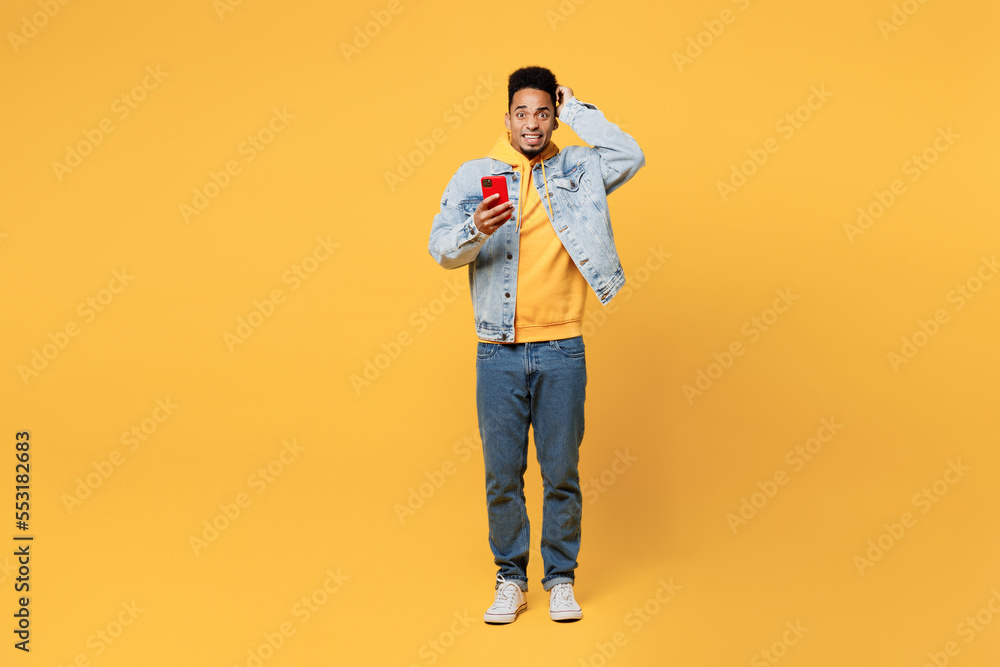 Fototapeta premium Full body sad shocked young man of African American ethnicity wear denim jacket hoody look camera hold head use mobile cell phone isolated on plain yellow background studio. People lifestyle concept.