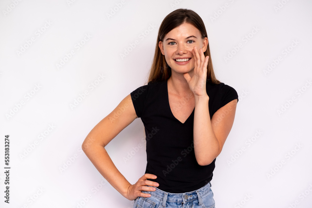 Portrait of happy young woman telling news covering mouth with hand over white background. Caucasian lady wearing black T-shirt and jeans whispering gossips. Secrecy concept Portrait of happy young woman telling news covering mouth with hand over white background. Caucasian lady wearing black T-shirt and jeans whispering gossips. Secrecy concept