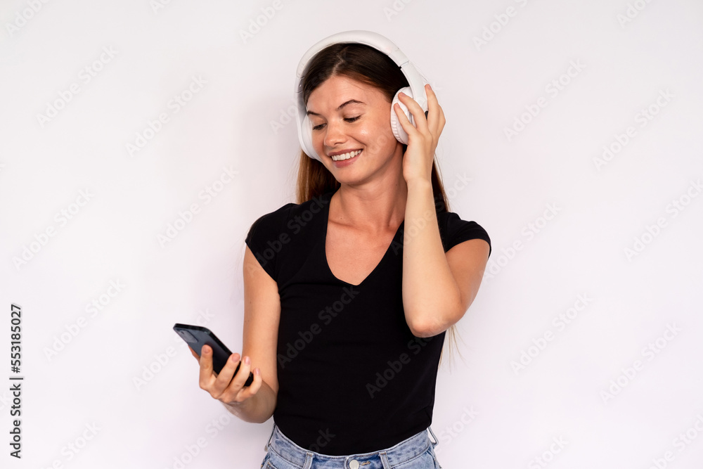 Portrait of happy young woman with mobile phone enjoying music in headphones. Caucasian lady wearing black T-shirt and jeans listening to music. Mobile technology concept Portrait of happy young woman with mobile phone enjoying music in headphones. Caucasian lady wearing black T-shirt and jeans listening to music. Mobile technology concept