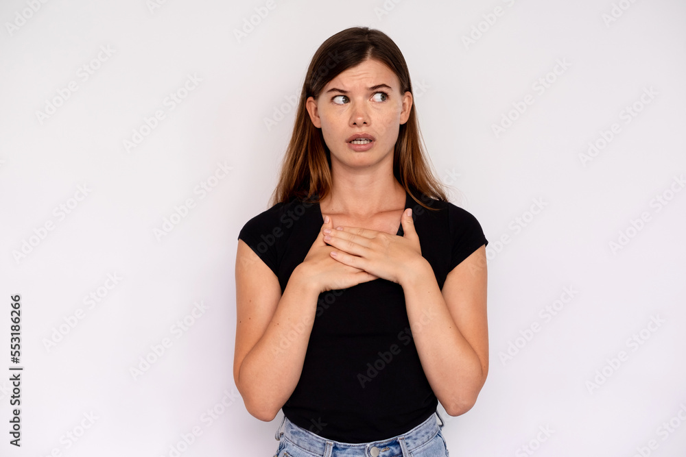 Portrait of displeased young woman looking away feeling guilt. Caucasian lady wearing black T-shirt and jeans touching chest in fear over white background. Anxiety concept