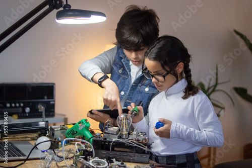 School children geek working on project on electronics. Girl in eyeglasses using solder, boy giving her advice. Education, hobby and robotics concept