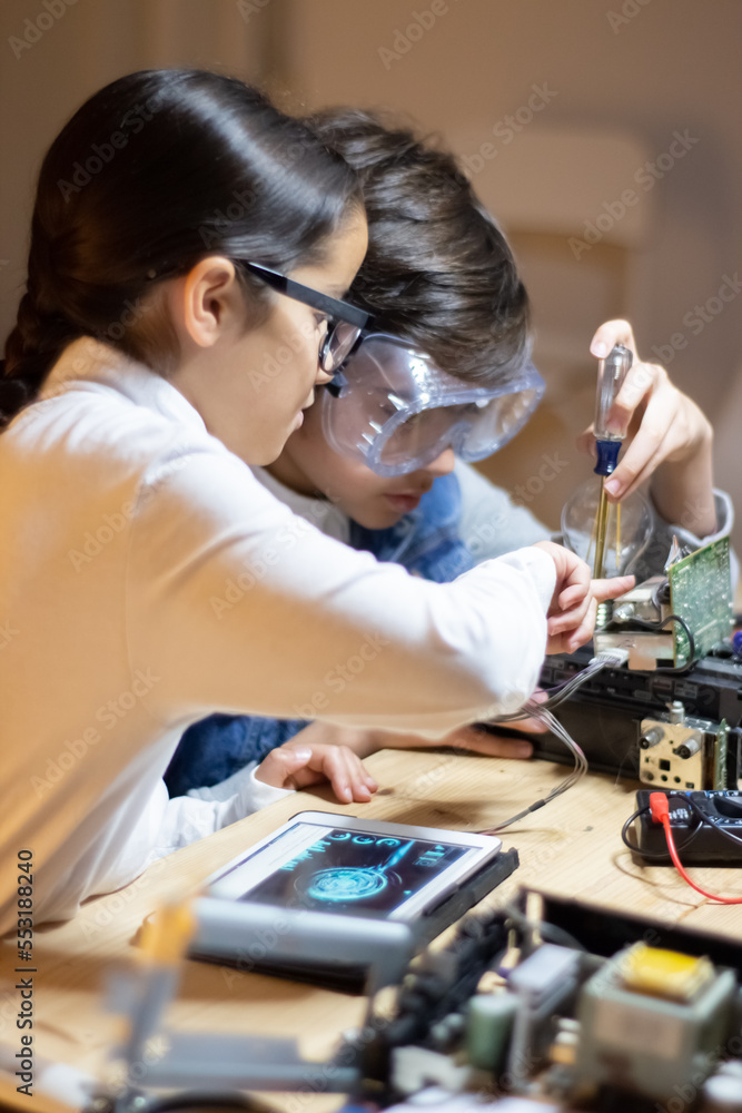 Focused school boy in protective glasses using screwdriver to fix elements in new electronic device. Girl helping him. Electronics, hobby concept