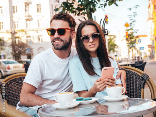 Smiling beautiful woman and her handsome boyfriend. Happy cheerful family. Couple drinking coffee in restaurant. They drinking tea at cafe in the street. Holding cup. Looking at smartphone, use apps