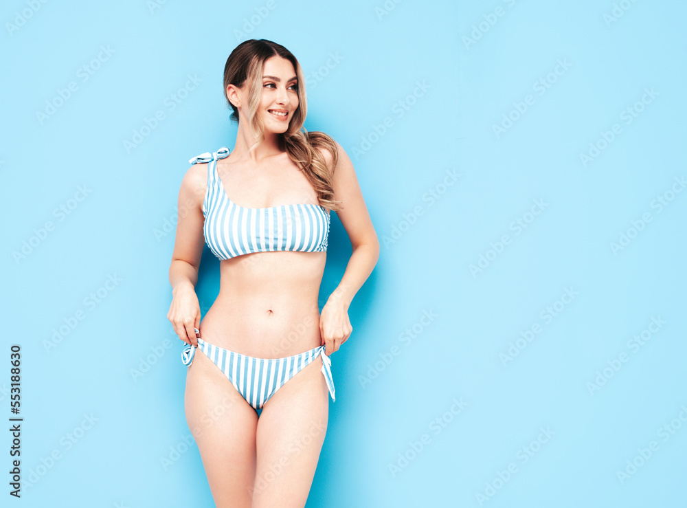 Naklejka premium Young smiling blond model in summer striped swimwear colourful bathing suit. Sexy carefree woman having fun and going crazy. Female posing near blue wall in studio. Cheerful and happy. Perfect body